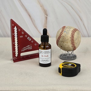 May include: A red protractor, a clear glass bottle with a dropper, a baseball on a stand, and a yellow tape measure on a white canvas background. The bottle label reads "Jarhead Aromatics The Sleuth Diffuser Refill".