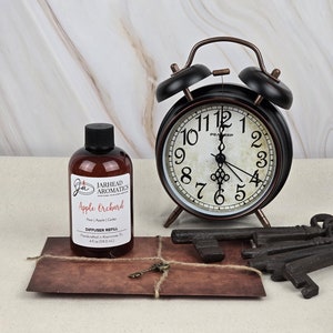 May include: A clear glass bottle with a brown label that reads "JARHEAD AROMATICS Apple Orchard Pear | Apple | Cedar DIFFUSER REFILL 4 fl oz (118.2 ml)". The bottle is sitting on a brown surface next to a vintage alarm clock, a key, and a brown envelope.