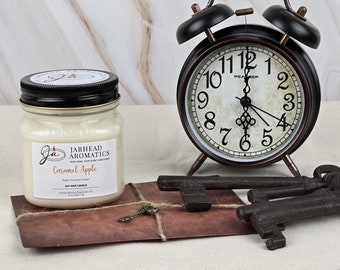 May include: A white soy wax candle in a glass jar with a black lid. The candle is labeled "Jarhead Aromatics" and "Caramel Apple". The candle is sitting on a brown envelope with a key and twine. A vintage alarm clock and keys are in the background.