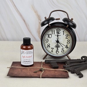 May include: A black and bronze alarm clock with a cream-colored face, next to a bottle of "Apple Orchard" diffuser refill. A small, antique key and a bundle of old keys are also present. The clock is approximately 6 inches tall.