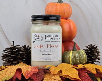 May include: A beige soy wax candle in a glass jar with a black lid. The candle is labeled "Jarhead Aromatics" and "Campfire Memories". The label also lists the scent notes as "Clove | Patchouli | Smoke". The candle is surrounded by fall-themed decorations, including pumpkins and maple leaves.