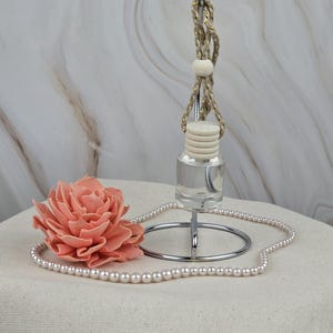 May include: A clear glass bottle with a white wooden cap and a beige rope hanging from it. The bottle is on a silver metal stand with a pearl necklace wrapped around it. A pink fabric flower is next to the necklace.