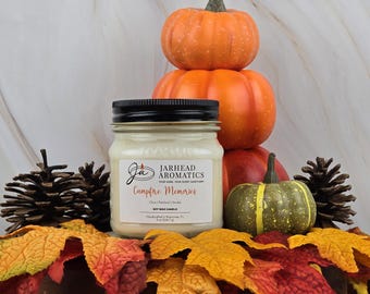 May include: A soy wax candle in a glass jar with a black lid. The candle is labeled "Campfire Memories" and is surrounded by fall-themed decorations, including pumpkins and pine cones.