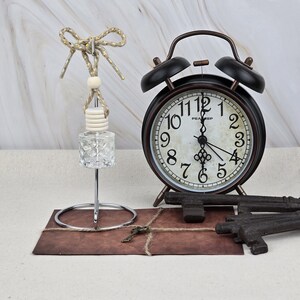 May include: A clear glass diffuser bottle with a wooden cap and gold-flecked cord, a black twin-bell alarm clock with a white face and black numerals, and two dark metal skeleton keys resting on a brown rectangular card tied with twine.