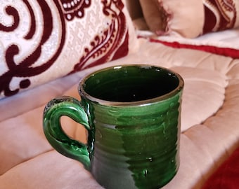 May include: A green ceramic mug with a handle. The mug is sitting on a white and red patterned surface.