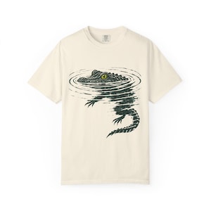 Op de afbeelding: Een crèmekleurig t-shirt met korte mouwen en een donkergroene grafische illustratie van een alligator die in het water zwemt. De alligator is afgebeeld met één zichtbaar geel oog en het water toont concentrische cirkelvormige rimpelingen.