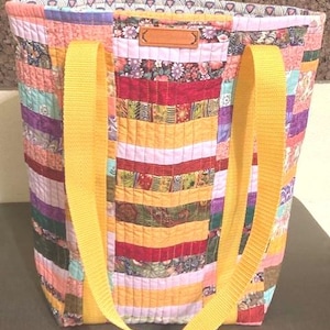 May include: A large, quilted tote bag with yellow straps. The bag is made of patchwork fabric in various colors, including pink, purple, red, green, and yellow. The bag has a brown leather tag with the word "Handmade" on it.