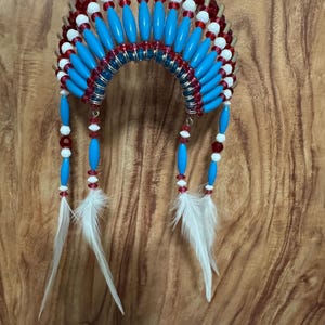 May include: A decorative Native American headdress featuring a curved, beaded design. The headdress is adorned with red, white, and blue beads, along with white feathers hanging from the bottom. The headdress is suspended by a gold string.