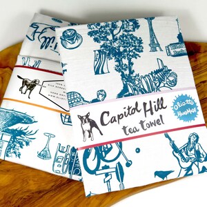 Puede incluir: Toalla de té blanca con un diseño azul y blanco que presenta puntos de referencia y escenas de Capitol Hill, Seattle. El texto "Capitol Hill tea towel" y "olio.to Handmade" están impresos en la toalla.