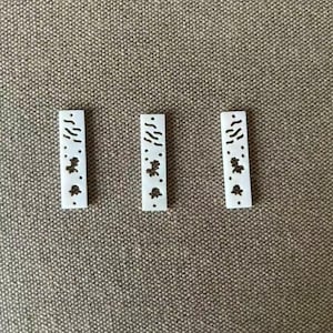 Puede incluir: Tres botones rectangulares blancos, cada uno con una serie de pequeñas marcas grabadas de color marrón oscuro. El diseño de cada botón consiste en un grupo de puntos y líneas finas y cortas dispuestas en un patrón vertical en el centro.