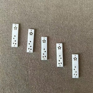 Puede incluir: Cinco colgantes rectangulares de concha blanca dispuestos en línea diagonal. Cada colgante presenta un pequeño orificio en la parte superior y una serie de formas recortadas, incluyendo una flor de cinco pétalos y varios pétalos pequeños.