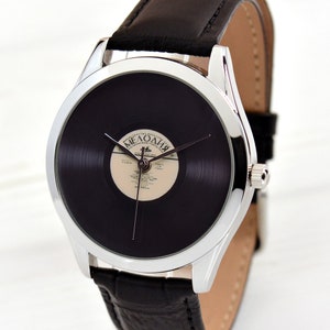 Puede incluir: Un reloj plateado con una correa de cuero negro. La esfera del reloj es un disco de vinilo negro con una etiqueta blanca que dice "Мелодия".