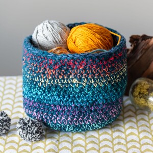 May include: A crocheted basket with a rainbow pattern, filled with balls of yarn in shades of grey, yellow, and brown. The basket is sitting on a table with a patterned tablecloth.