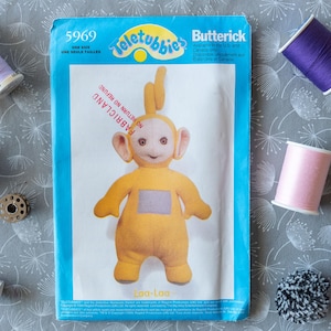 Pode incluir: Um padrão de costura azul e branco com um personagem amarelo dos Teletubbies chamado La-La. O padrão é etiquetado como "5969" e "Teletubbies" com o logotipo da Butterick. O padrão é para um brinquedo de pelúcia.