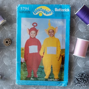Pode incluir: Um padrão de costura Butterick para fantasias de Teletubbies, com Po e Laa-Laa, com uma borda azul. A imagem inclui carretéis de linha em várias cores, incluindo roxo, rosa e azul, em um fundo de tecido cinza estampado.