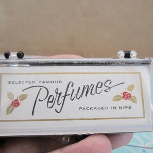 May include: A rectangular clear plastic case with a hinged lid containing a white insert. The insert features the text "SELECTED FAMOUS Perfumes PACKAGED IN NIPS" in black, flanked by two small illustrations of red roses with gold leaves.