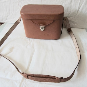 May include: A structured, rectangular brown leather camera bag with a textured finish. The bag features a top lid with a flap closure secured by a silver-toned metal latch. It includes a long, adjustable shoulder strap with a matching leather pad.