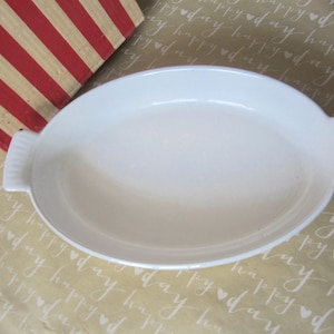 May include: A white oval baking dish with handles. The dish is made of ceramic and has a smooth, glossy finish. The dish is empty and ready for use. A red and white striped box is in the background.
