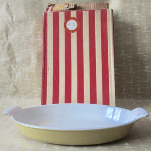 May include: A yellow oval baking dish with white interior and handles. Behind it is a vintage box with red and cream vertical stripes and a round sticker. The background is a light brown surface.