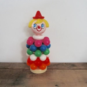 May include: A vintage plastic clown toy with a red hat, yellow hair, and a smiling face. The clown's body is made of stacked colorful balls in red, orange, white, green, blue, and pink. The toy is on a wooden surface.