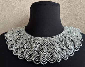 May include: A silver beaded collar with a scalloped edge. The intricate openwork design is displayed on a black mannequin. The collar is a statement piece, perfect for adding a touch of elegance to any outfit.
