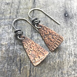 Könnte beinhalten: Ein Paar kupferne Dreiecksohrringe mit strukturierter Oberfläche und silbernen Haken. Die Ohrringe haben ein geometrisches Muster.