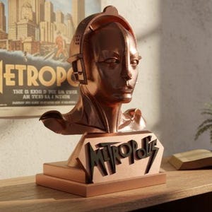 May include: A copper-colored bust of the Maschinenmensch robot from the film "Metropolis" sits on a wooden surface. The bust is mounted on a base with the film's title. A vintage movie poster is in the background.