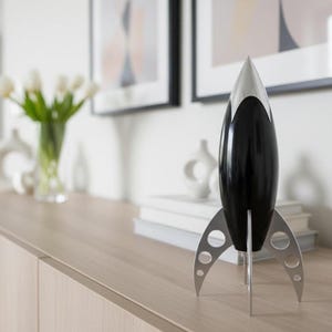 May include: A black and silver rocket ship decorative object. The rocket has a silver tip and fins, with a black body. The rocket is standing on a wooden surface. A vase of white flowers and framed art are in the background.
