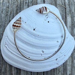 May include: A silver-toned bracelet featuring two small seashells with brown and white stripes. The bracelet is set against a large, white seashell, resting on a weathered wooden surface. The seashells are attached to the bracelet with a clear adhesive.