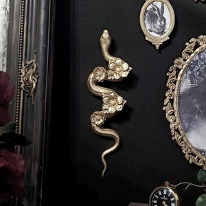 May include: A gold-coloured wall sculpture of a snake with three floral accents along its body. The snake is depicted in a winding, S-shaped pose with a textured surface, mounted on a dark wall alongside various framed pictures and a small clock.