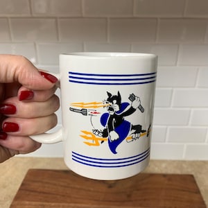 May include: White ceramic mug with a cartoon cat graphic. The cat is black with a blue cape and is holding a weapon. The mug has two blue stripes near the top and is held by a person.