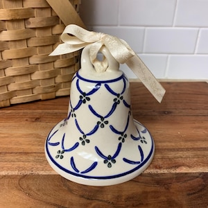 May include: A ceramic bell with a cream-colored base and a blue and green floral design. The bell has a cream-colored ribbon tied at the top. The bell is sitting on a wooden surface.