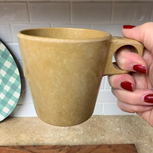 May include: A tan-colored mug with a unique handle design. The mug is held by a hand with red nail polish. The mug has a cylindrical shape and a textured surface. The mug is made of a durable material.