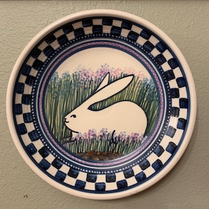 May include: A round ceramic plate with a white rabbit illustration. The plate features a blue and white checkered border, a purple and blue floral design, and a white rabbit in a field of flowers.