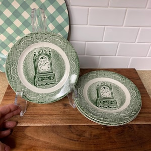 May include: Set of green and white decorative plates with a clock design. The plates have a white center with a green clock illustration and a green patterned border. The plates are displayed on a wooden surface.