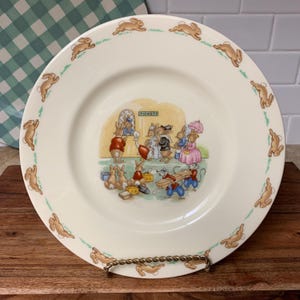 May include: A cream-colored ceramic plate with a whimsical illustration of rabbits. The plate features a border of running rabbits and a central scene of rabbits at a ticket booth. The word "TICKETS" is visible above the booth.