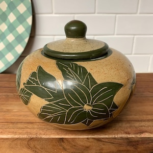 May include: A ceramic jar with a lid, featuring a floral design. The jar is tan with dark green floral patterns and a dark green lid. The jar is sitting on a wooden surface.