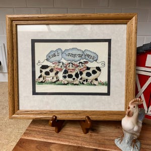 May include: Framed cross-stitch artwork featuring three cows with black and white spots. The cows are positioned on a green grassy area, with the text "Home is Where the Herd Is" above them. The frame is made of light brown wood.