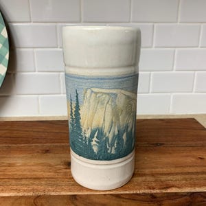 May include: A white ceramic tumbler with a landscape design. The design features a mountain, trees, and a blue sky. The tumbler is cylindrical and stands on a wooden surface.