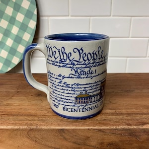 May include: A ceramic mug with a blue handle and rim, featuring the text "We the People" in a script font. The mug also displays text from the U.S. Constitution and an illustration of a building, with "1787 Bicentennial of" printed at the bottom.
