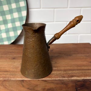 May include: A hammered copper coffee pot with a tapered body, a wide pouring spout, and a long, turned wooden handle attached with a metal bracket. The metal surface features a textured, dimpled finish with a warm, aged brown patina.