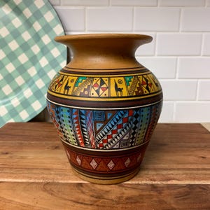 May include: A decorative ceramic vase with a wide base and a narrow neck. The vase features a tan body with colorful geometric patterns in yellow, blue, red, and brown. The design includes llama figures and various shapes. The vase is displayed on a wooden surface.