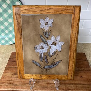May include: A framed floral art piece featuring white lilies with brown centers and gray-green leaves. The artwork is set against a tan background and framed in a light brown wooden frame. The frame is supported by two clear plastic stands.