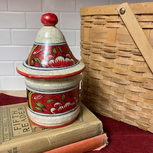 May include: A ceramic jar with a lid, featuring a white base with red and green floral designs. The jar has a crackled glaze and a red knob on the lid.