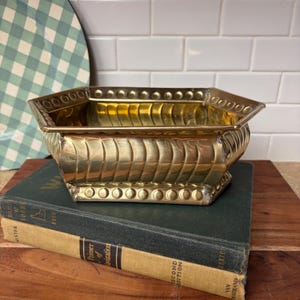 May include: A hexagonal brass bowl featuring a polished metallic finish, a scalloped vertical ribbed pattern along the sides, and a decorative border of raised circular studs along both the top rim and the base of the vessel.