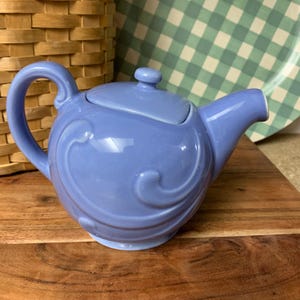 May include: A periwinkle blue ceramic teapot with a curved handle and spout. The teapot features a lid with a small knob and decorative swirl designs on the body. A wicker basket and a green and white checkered plate are in the background.