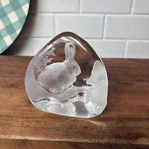 May include: A clear glass paperweight with a triangular shape featuring an etched, frosted design of a rabbit sitting on a branch. The interior of the glass displays a detailed, textured silhouette of the animal surrounded by stylized foliage.