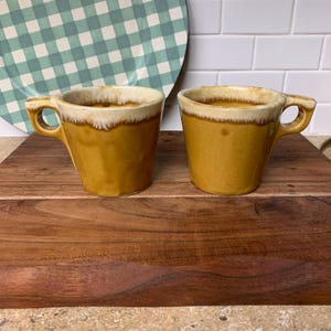 May include: Two vintage mustard-yellow coffee mugs with a cream-colored rim and a unique handle design. The mugs are displayed on a wooden cutting board, with a green and white checkered plate in the background. The mugs have a retro aesthetic.