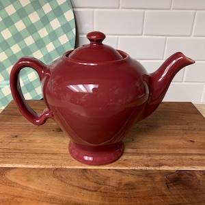 May include: A burgundy ceramic teapot with a matching lid and handle. The teapot has a rounded body, a spout, and sits on a small base. The teapot is displayed on a wooden cutting board.