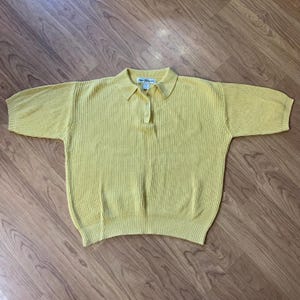 May include: A pale yellow knit polo shirt with short sleeves. The shirt has a collared neckline and a three-button placket. The fabric has a ribbed texture. The shirt is laying flat on a wooden surface.
